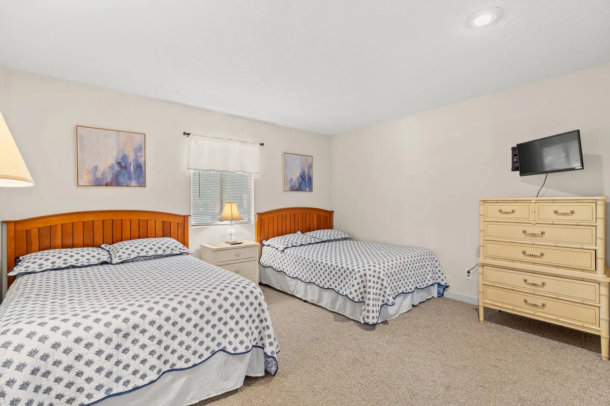 Depicts a second bedroom with two inviting beds, a nightstand with a lamp, a dresser, and decorative wall art. The mood is welcoming and designed for shared moments, ideal for families or friends.