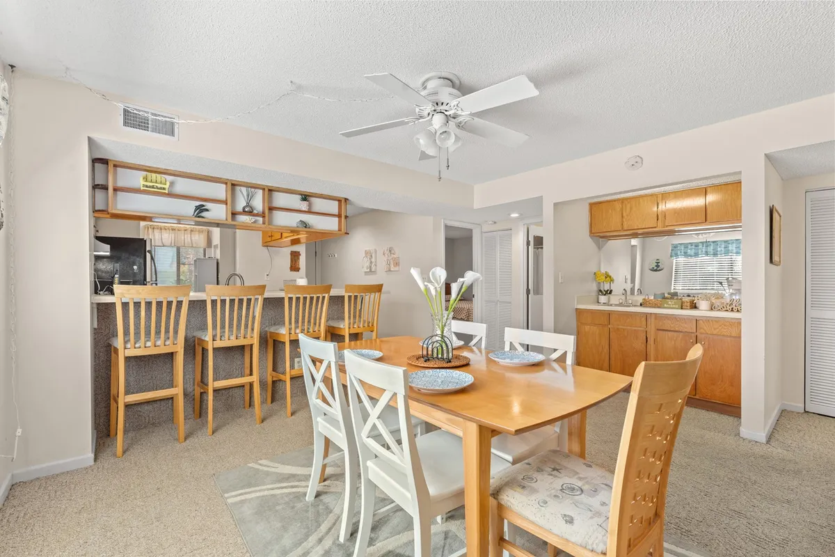 This image focuses on the dining area, featuring a spacious dining table with bar stools and a glimpse of the kitchen in the background. Decorative plants add a touch of warmth, while ample seating ensures everyone has a place at the table for shared meals or celebrations.