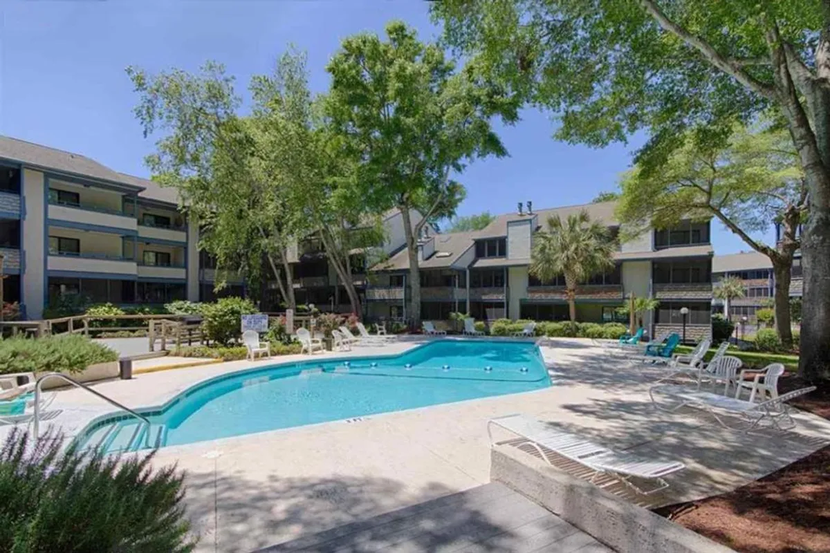 A vibrant exterior pool scene framed by lush greenery, with sun-soaked decks, inviting swimming pool, and balconies visible in the background. The mood is cheerful, with space for swimming, sunbathing, and casual gatherings.