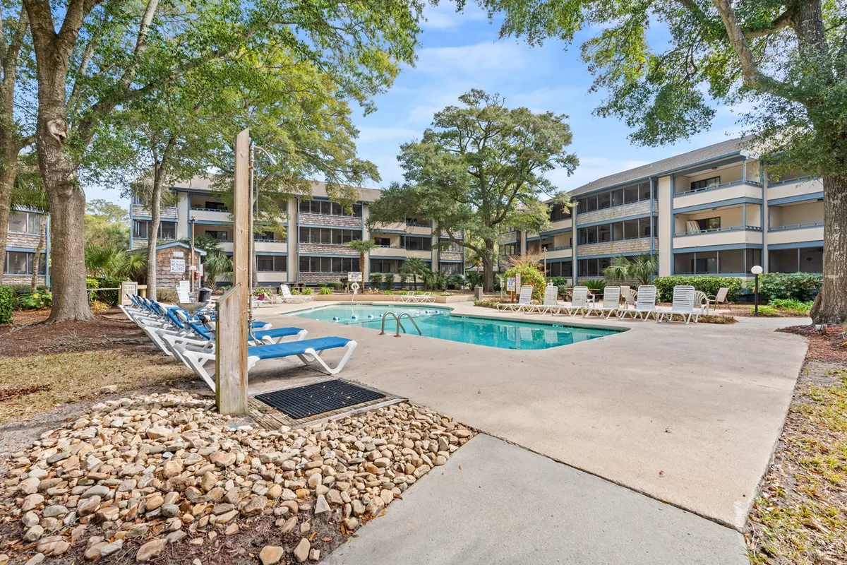 The image showcases the inviting outdoor pool area of Summertree Village A5, surrounded by lush trees and lounge chairs, with the condo building facade in view. The setting suggests a tranquil spot for swimming, sunbathing, and socializing, all just a short stroll from the beach.