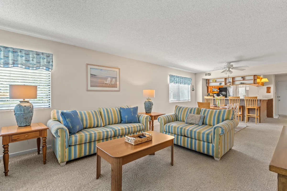 Shows a living room with striped sofas (including a sleeper sofa), decorative vases, a wooden coffee table, framed artwork, and a bar area. The arrangement is perfect for relaxing or hosting guests, reflecting comfort and togetherness.