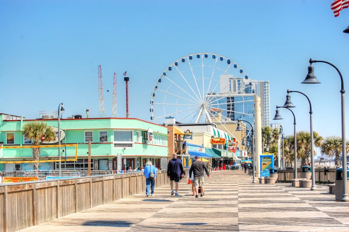 This vibrant boardwalk scene features a ferris wheel, colorful storefronts, and groups of people strolling and enjoying various attractions. The lively atmosphere conveys a sense of fun and adventure, perfect for multi-generational outings.