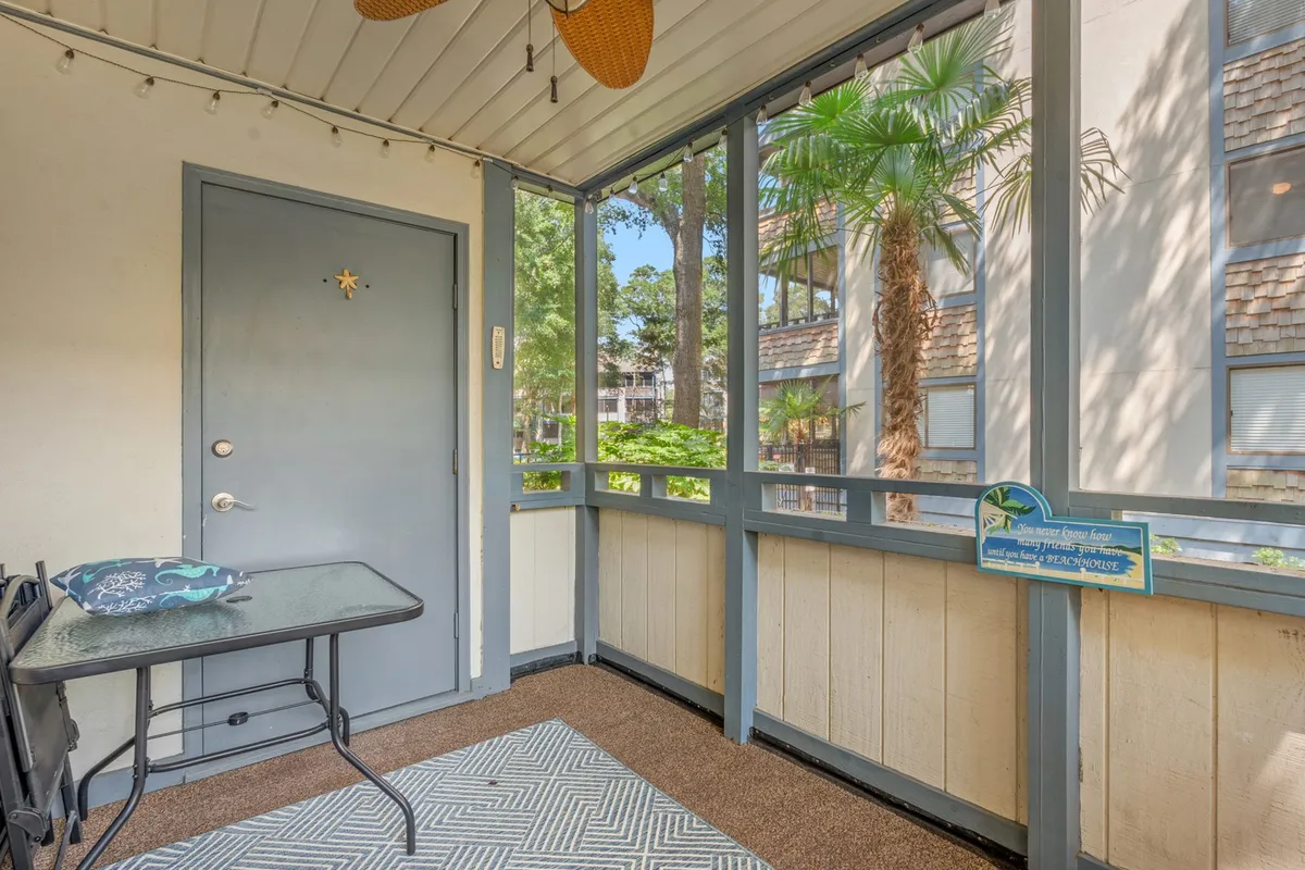A cozy screened porch with a table and chairs set atop a decorative rug, featuring window views of lush palm trees and a door with a charming star motif. The ambiance is tranquil, offering the perfect setting for morning coffee or peaceful relaxation.