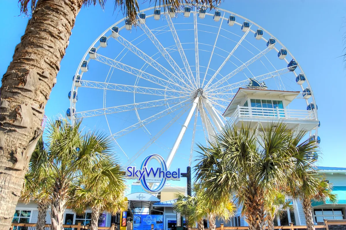 The iconic Myrtle Beach SkyWheel rises above the surrounding palm trees and the vibrant entrance plaza, inviting families to experience panoramic views and thrilling rides. The sunny sky and bustling energy capture the excitement and attractions nearby.