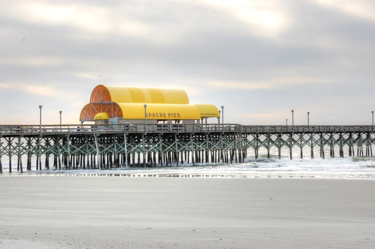 An inviting view of Apache Pier stretches over rolling ocean waves, with its distinctive yellow roof creating a focal point. The tranquil beachfront scene hints at both daytime outings and peaceful sunset strolls.