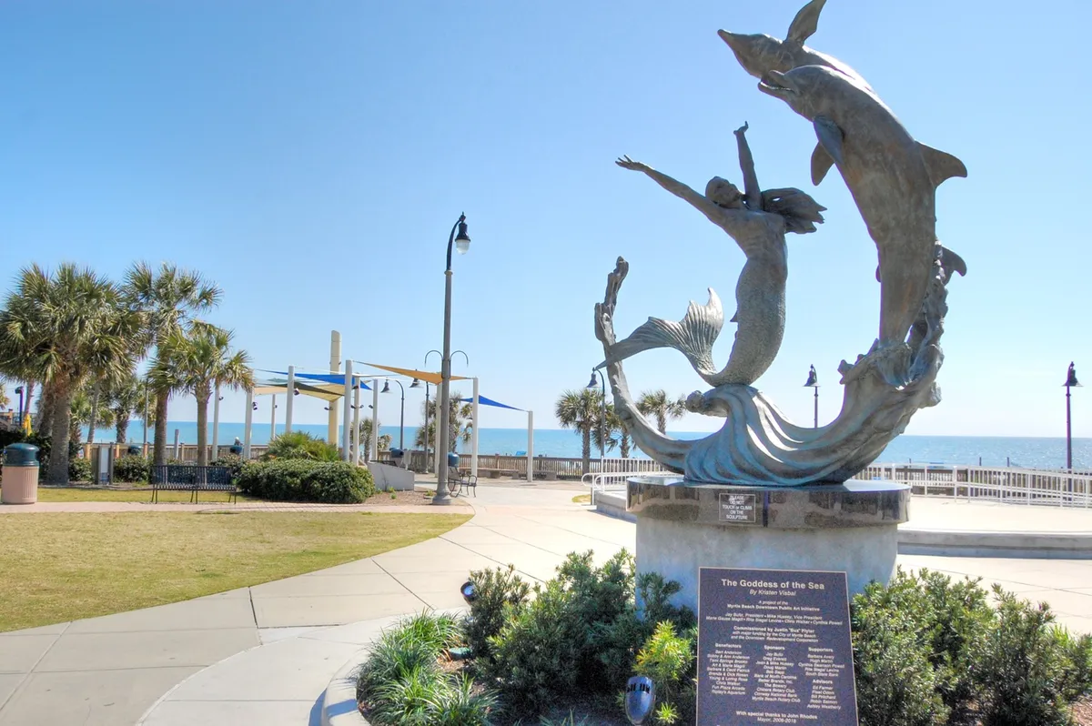 A striking sculpture of the Goddess of the Sea sits along a scenic oceanfront path with palm trees and the sea in the background. Visitors are seen strolling and relaxing, emphasizing the inviting and picturesque coastal atmosphere.